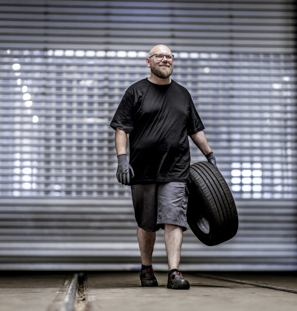 Handwerker in schwarzem T-Shirt und grauen Shorts trägt einen Reifen in einer Werkstatt.