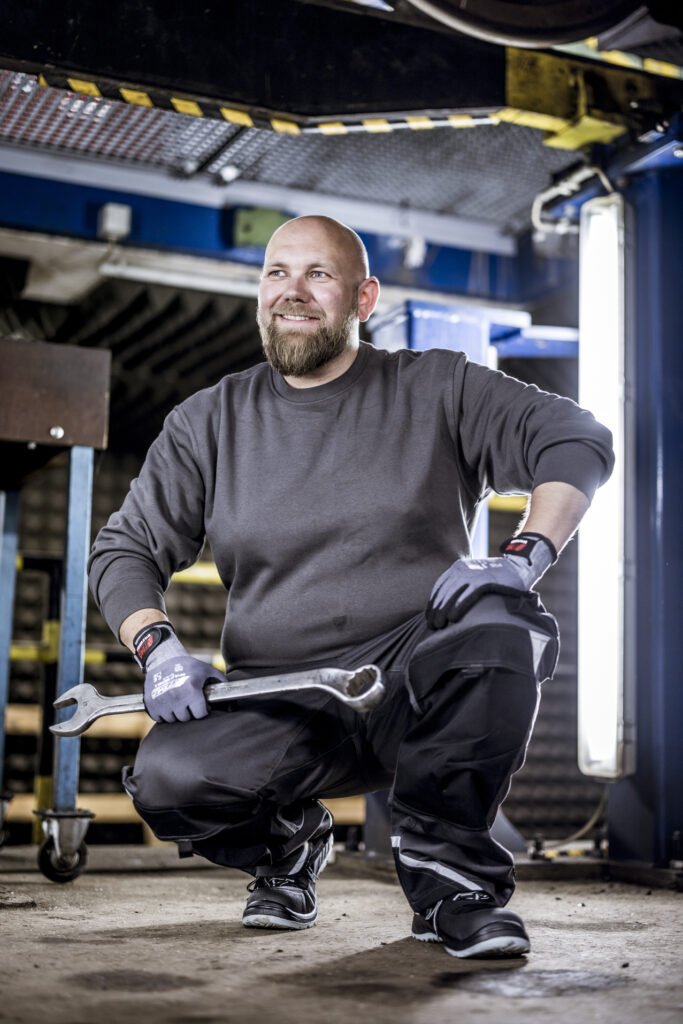 Mechaniker in schwarzer Arbeitskleidung kniet mit einem Schraubenschlüssel in der Hand in einer Werkstatt.