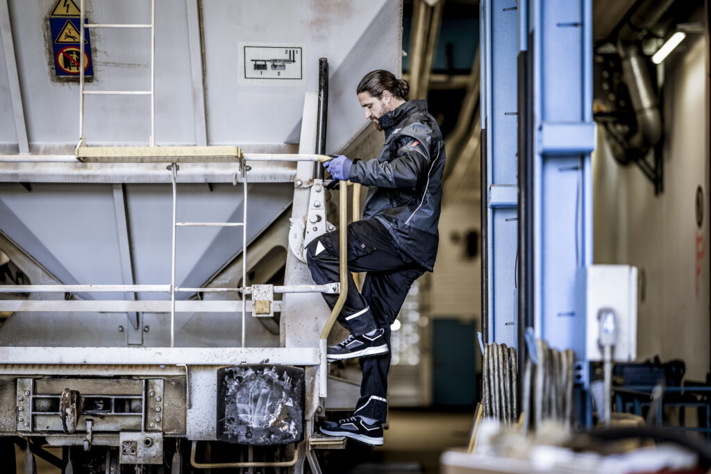 Handwerker in schwarzer Arbeitsjacke und Schutzkleidung steigt auf eine Industriemaschine und arbeitet mit Handschuhen.