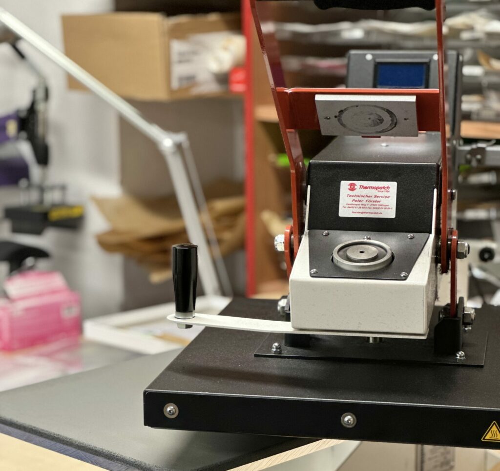 Thermopatch Transferpresse mit Hebelmechanismus und digitaler Steuerung für den professionellen Textildruck, auf einer stabilen Werkbank positioniert.