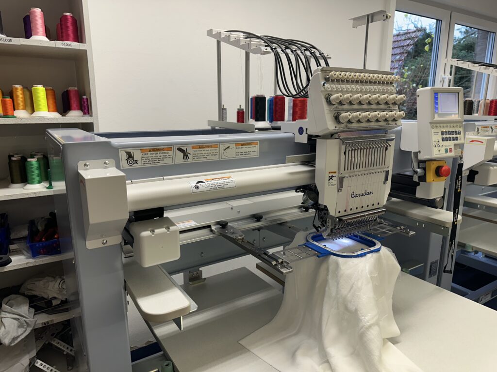 Professionelle Mehrkopf-Stickmaschine von Barudan bei der Arbeit in der Stickerei-Werkstatt, mit eingespanntem Stoff und Garnregal im Hintergrund.