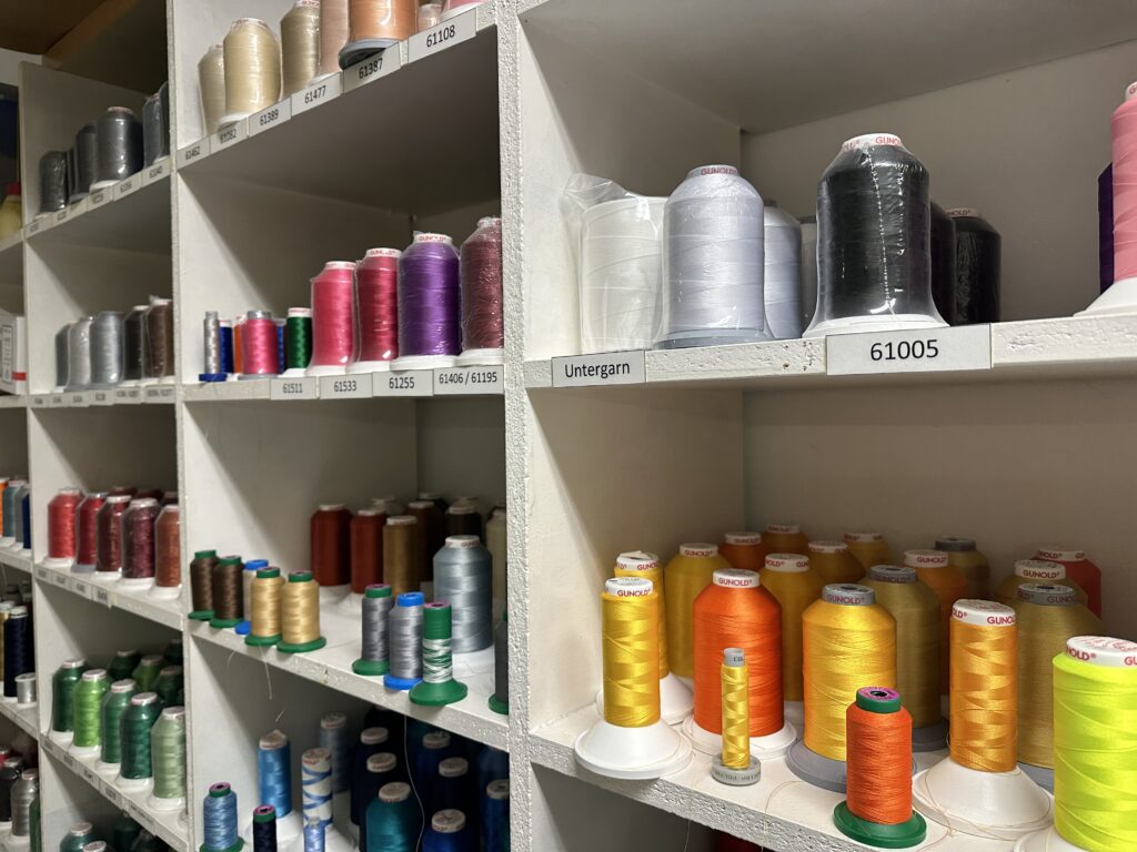 Sortierte Garnrollen in verschiedenen Farben in einem Regalsystem, beschriftet mit Farbnummern und Bezeichnungen.