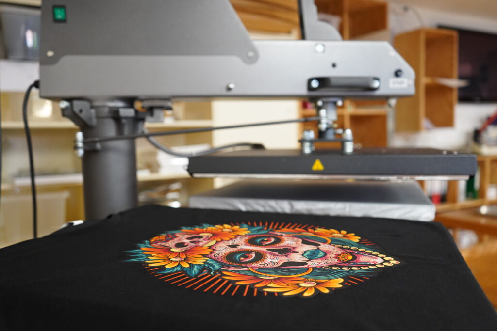 Schwarzes T-Shirt mit farbenfrohem Totenkopf-Motiv unter einer geöffneten Textilpresse, im Hintergrund Regale mit Materialien.