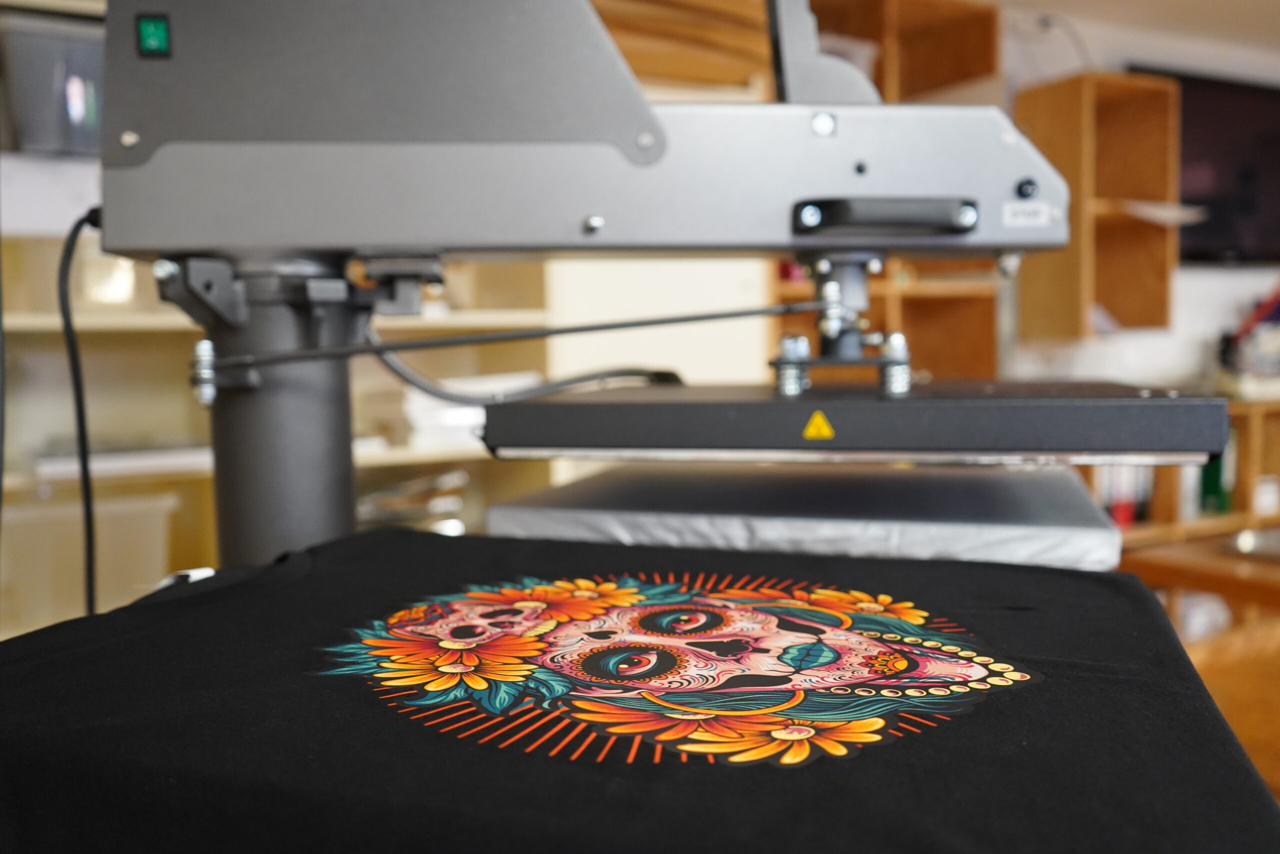 Schwarzes T-Shirt mit farbenfrohem Totenkopf-Motiv unter einer geöffneten Textilpresse, im Hintergrund Regale mit Materialien.