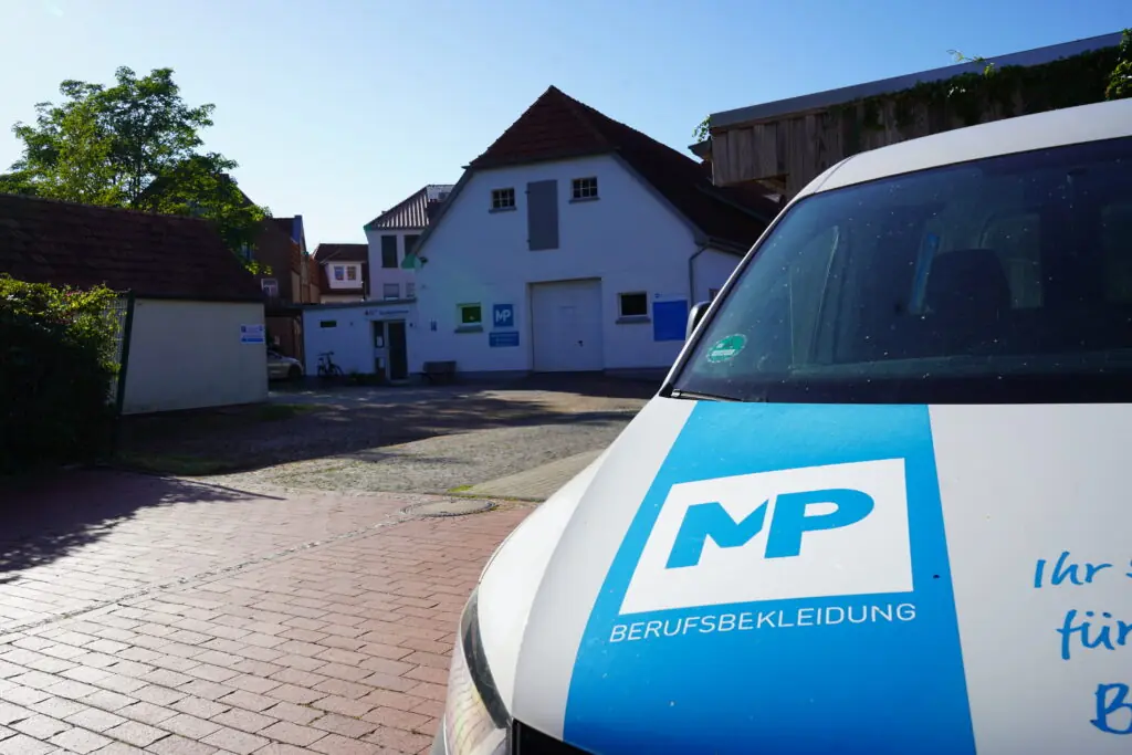 Gebäude und Fahrzeug mit MP-Logo für Berufsbekleidung in einem Innenhof bei Sonnenschein.