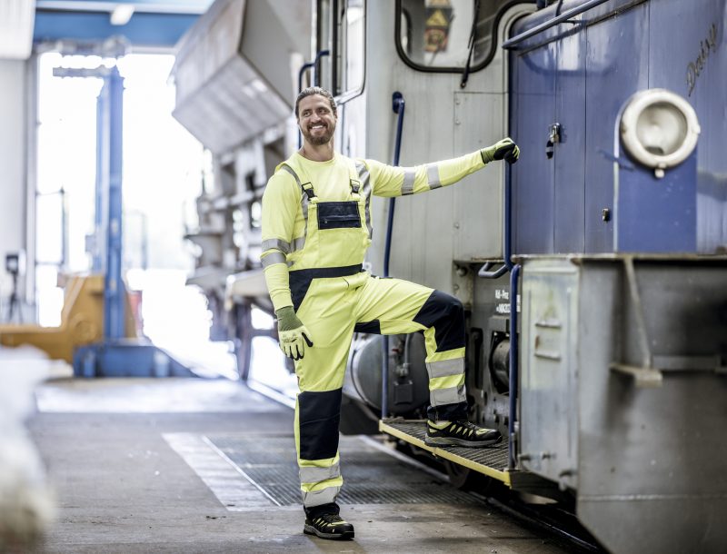 Arbeiter in neongelber Warnschutzkleidung mit reflektierenden Streifen steigt in eine Lokomotive in einer Werkstatthalle.