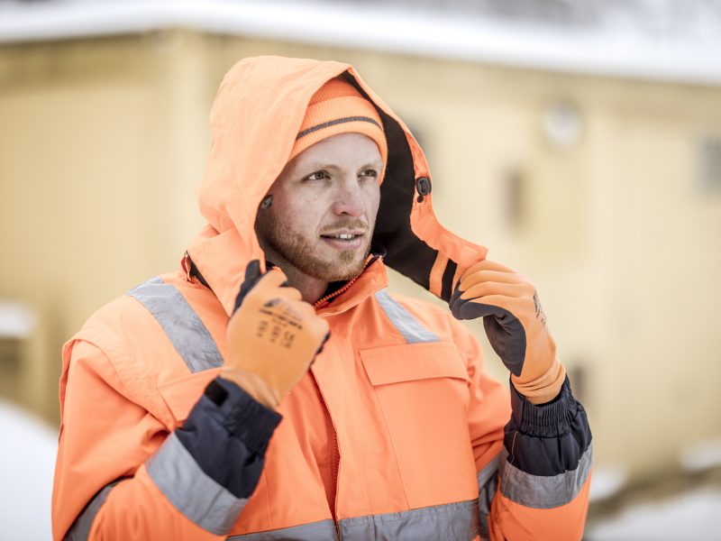 Arbeiter in oranger Warnschutzjacke mit reflektierenden Streifen setzt seine Kapuze auf, während er sich im Schnee aufhält.