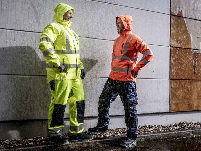 Zwei Arbeiter in wasserdichter Warnschutzkleidung stehen im Regen und lachen, einer in neongelber, der andere in orangefarbener Jacke mit reflektierenden Streifen.
