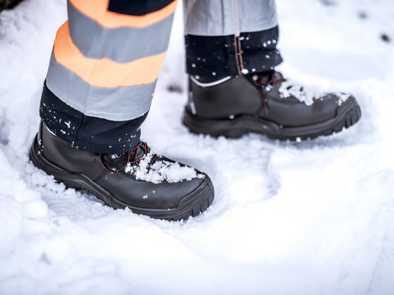 Sicherheitsschuhe mit rutschfester Sohle im Schnee, getragen mit reflektierender Warnschutz-Arbeitshose.