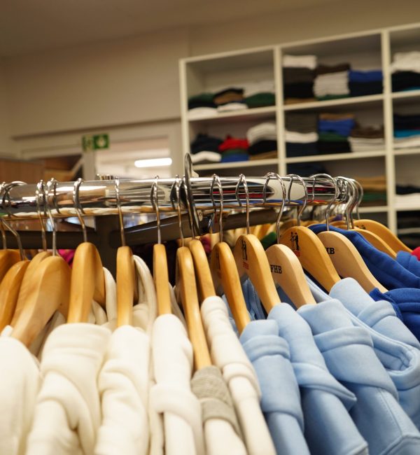 Nahaufnahme eines runden Kleiderständers mit farblich sortierten Hoodies auf Holzbügeln; im Hintergrund gefaltete Kleidung in Regalen.