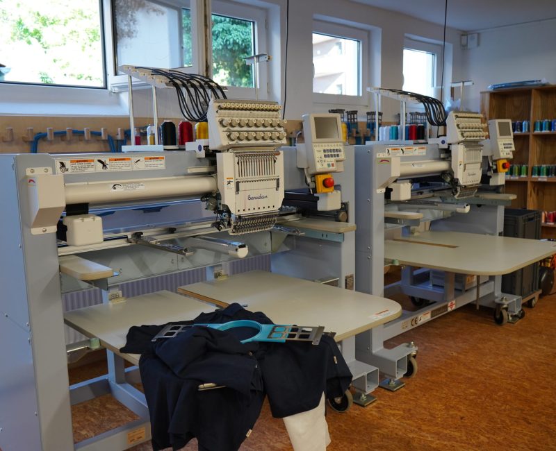 Drei professionelle Stickmaschinen in heller Werkstatt, im Vordergrund ein eingespanntes Kleidungsstück, im Hintergrund Garnrollenregale.