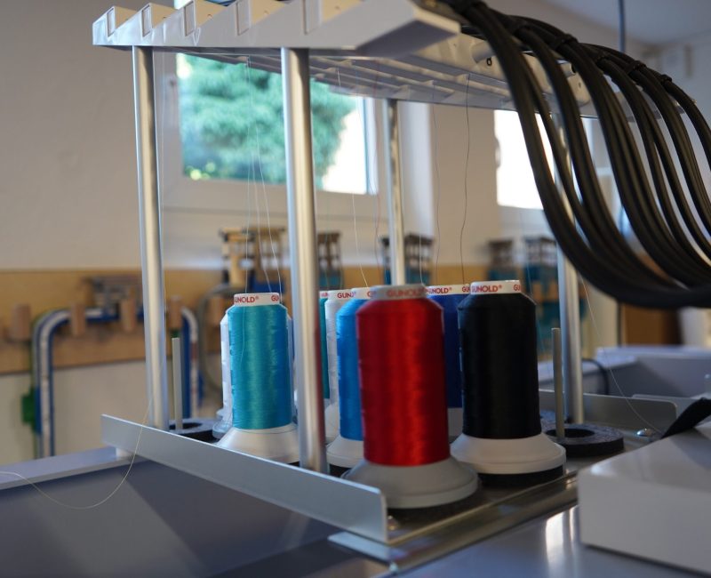 Mehrere Garnrollen in verschiedenen Farben sind an einer Stickmaschine eingespannt, darüber führen schwarze Kabel zu den Nadelköpfen.
