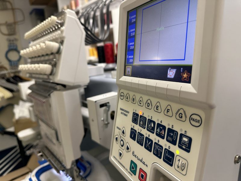 Nahaufnahme einer professionellen Barudan-Stickmaschine mit digitalem Display und mehreren Nadelköpfen in einer Stickerei-Werkstatt.