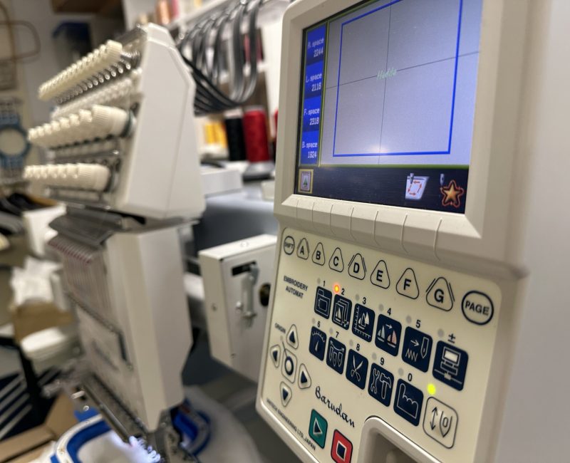 Nahaufnahme einer professionellen Barudan-Stickmaschine mit digitalem Display und mehreren Nadelköpfen in einer Stickerei-Werkstatt.